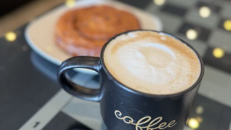 A cappuccino in a black mug and a spiral pastry on a chessboard table